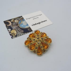 Puede incluir: Un broche dorado de forma cuadrada con un diseño de piedras de imitación naranja y marrón. El broche está engastado en un patrón floral.