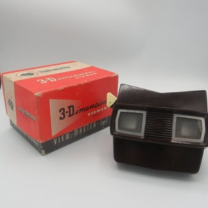 May include: A vintage View-Master 3-D viewer in brown plastic with a white knob. The viewer is in its original box, which is red with white and black lettering. The box reads "View-Master 3-D Viewer Model 1".