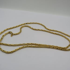 May include: A long, gold-toned necklace with a rectangular link chain design. The necklace is laid out on a white surface, showcasing its length and the details of the chain links. The clasp is visible.