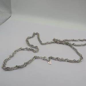 May include: A silver-coloured necklace with a twisted rope design. The necklace is lying on a white surface. The necklace has a clasp and a small leaf-shaped charm.
