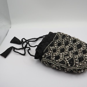 May include: A black beaded drawstring pouch with a scalloped design. The pouch has a black satin lining and a drawstring closure with black tassels. It is decorated with silver-coloured beads and rhinestones.