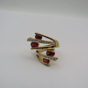May include: A gold-toned ring with a unique design. The ring features four oval-shaped, deep red gemstones and multiple rows of small, clear gemstones. The ring's design incorporates a series of angled bars.