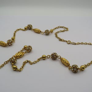 May include: A gold-toned necklace with a delicate chain and decorative elements. The necklace features textured oval beads, round crystal-studded spheres, and smaller round beads. The chain is connected with a clasp.