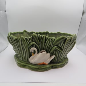 May include: A green ceramic planter with a swan design. The planter has a rectangular shape with a textured, leafy design around the edges. A white swan with an orange beak and feet is featured on the front. Suitable for indoor or outdoor use.