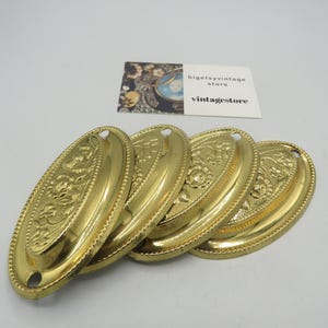May include: Four vintage brass oval drawer pulls with floral and beaded designs. Each pull has a small hole at the top for mounting. The pulls are arranged on a white surface. A business card is in the background.