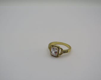 Op de afbeelding: Een goudkleurige ring met een rechthoekige, heldere edelsteen in het midden. De edelsteen is omgeven door een vierkant frame met kleinere, heldere stenen. De band van de ring heeft een textuur.