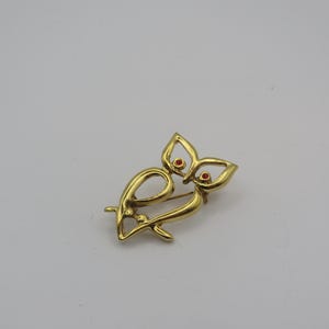 May include: A gold-coloured owl brooch with red detailing. The brooch has an openwork design with stylised wings and body, and small red stones for eyes. This vintage pin is a jewellery accessory.