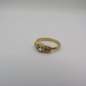 May include: A gold-toned ring with a large, clear, round gemstone at the center, flanked by smaller, clear accent stones. The ring has a smooth, polished band and a decorative setting for the gemstones.