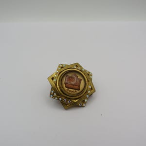 May include: A gold-toned brooch with a central circular design featuring a brown image and the word "ROMA". The brooch is surrounded by a starburst design with clear crystal accents.