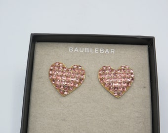 Op de afbeelding: Een paar hartvormige oorbellen met roze kristallen accenten. De oorbellen zijn verpakt in een zwarte doos met de merknaam "BAUBLEBAR" erop gedrukt.