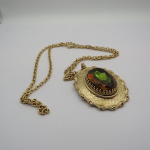May include: Gold-tone necklace with an oval pendant. The pendant features a large, multi-colored gemstone in shades of green and orange, set within a textured, ornate frame. The chain is a delicate link style.