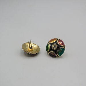 May include: A pair of gold-tone clip-on earrings with a black background and colourful enamel accents. The earrings feature a round shape with a raised design of five oval-shaped stones in shades of green, orange, red, and purple. A small clear rhinestone is centred in the design.