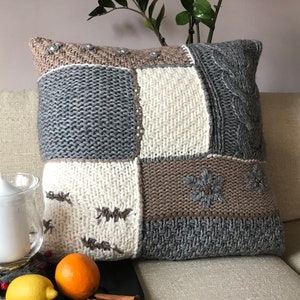 May include: Cozy knitted throw pillow in grey, beige, and off-white hues. The patchwork design features various knit textures, including cable knit and embroidered snowflake patterns.  The pillow is square-shaped and rests on a beige sofa.  It's a perfect addition to a rustic or farmhouse-style living room decor.