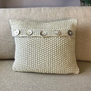 May include: A beige knitted throw pillow with a row of silver buttons across the front.