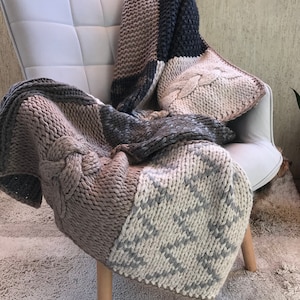 Può includere: Una coperta di maglia con un design a patchwork in tonalità di marrone, grigio e bianco. La coperta è drappeggiata su una sedia.