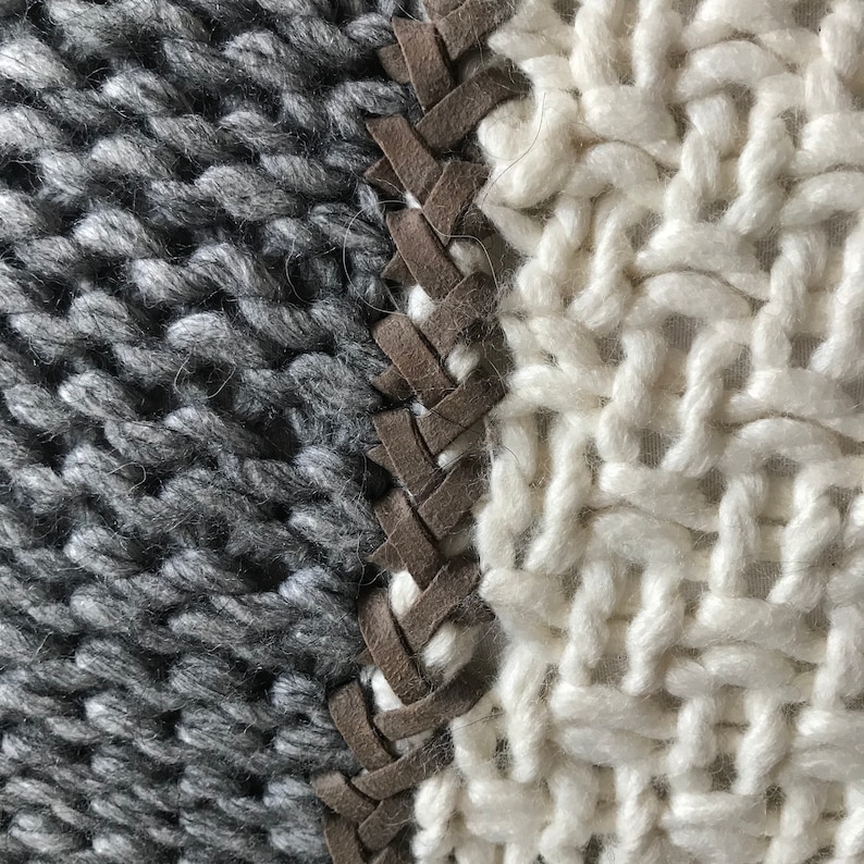 May include: Close-up of a knitted textile featuring a striking contrast of textures and colors. One section showcases a chunky, dark gray knit, while the adjacent section displays a lighter, off-white knit with a distinct open weave. A brown leather-like braid acts as a striking seam between the two knitted fabrics, adding a unique design element. The image highlights the contrast in knitting styles and the incorporation of leather detailing.