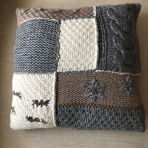 May include: A knitted throw pillow with a patchwork design. The pillow features various shades of gray, brown, and off-white knit fabric in different textures, including cable knit and embroidered floral accents. The overall style is cozy and rustic.