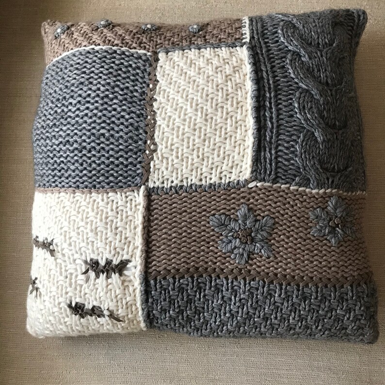 May include: A knitted throw pillow with a patchwork design. The pillow features various shades of gray, brown, and off-white knit fabric in different textures, including cable knit and embroidered floral accents. The overall style is cozy and rustic.