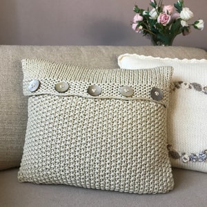 May include: A beige knitted pillow with a row of white buttons across the top. The pillow is on a beige couch.