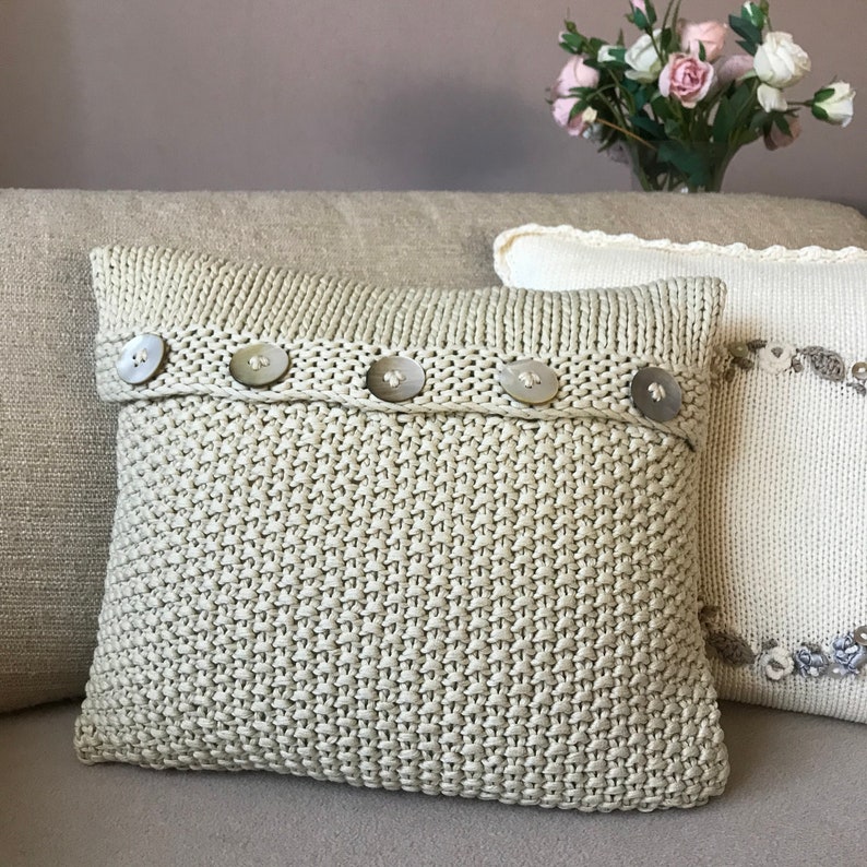 May include: A beige knitted pillow with a row of white buttons across the top. The pillow is on a beige couch.