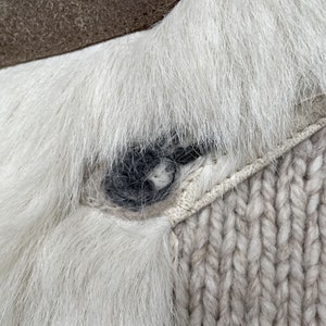 May include: Close-up of a section of a white fur item with a dark gray accent. The item is attached to a light gray, chunky knit fabric. The fur has a soft, textured appearance.