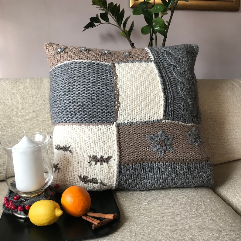 May include: Cozy knitted throw pillow in grey, beige, and off-white hues. The patchwork design features various knit textures, including cable knit and embroidered snowflake patterns.  The pillow is square-shaped and rests on a beige sofa.  It's a perfect addition to a rustic or farmhouse-style living room decor.