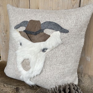 May include: A square, beige knitted pillow with a decorative goat face. The goat face features a brown hat, gray horns, and a white fur beard. The pillow is a cozy home decor item.
