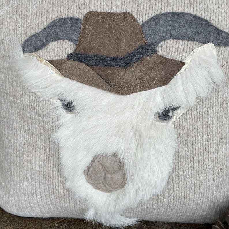 May include: A decorative pillow with a goat face design. The goat has a white fur face, gray horns, and a brown hat. The pillow has a light beige knitted background.