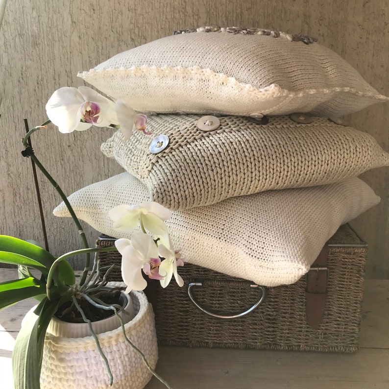 May include: A stack of three knitted throw pillows in cream and beige with decorative buttons. The pillows are sitting on a woven wicker storage box with a leather strap. A white orchid plant is in a white woven pot in the foreground.