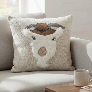 May include: A decorative throw pillow with a textured, light beige cover. The pillow features an applique of a goat's face, complete with a brown cowboy hat, gray horns, and a fluffy white beard. The pillow is on a light gray sofa.