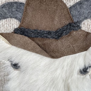 May include: Close-up of a decorative item featuring a brown suede hat with a dark gray band, gray felt horns, and a white fur base. The background is a cream-colored knitted fabric.