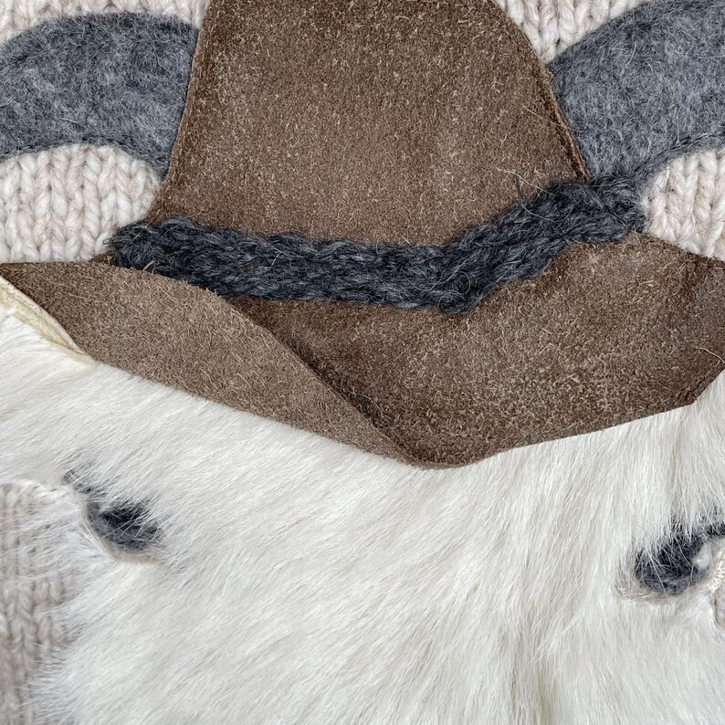 May include: Close-up of a decorative item featuring a brown suede hat with a dark gray band, gray felt horns, and a white fur base. The background is a cream-colored knitted fabric.