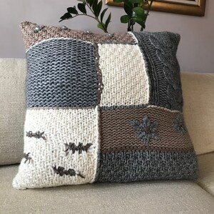 May include: A knitted patchwork throw pillow in shades of gray, beige, and off-white. The pillow features various knit textures, including cable knit and embroidered snowflake designs.  The square patches are sewn together to create a cozy, textured look. The pillow is perfect for adding a touch of rustic charm to any living space.