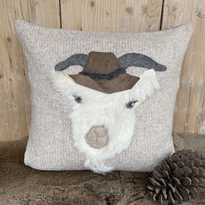 May include: A square, beige knit pillow featuring a goat face with a brown cowboy hat. The goat's face is made of white faux fur, with gray horns and eyes. A pinecone rests on the right side of the pillow.