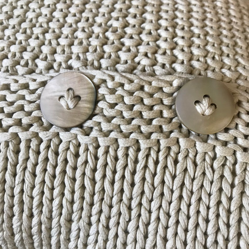 May include: Close-up of a beige knitted fabric with two round, light brown buttons. The buttons have a small heart-shaped design in the center.