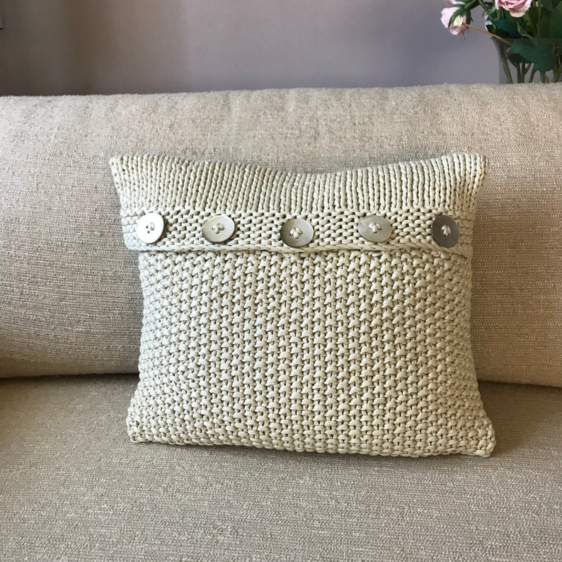 May include: A beige knitted throw pillow with a row of silver buttons across the front.