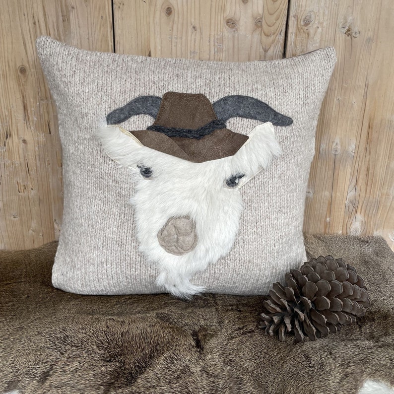 May include: A decorative square pillow with a goat face design. The pillow features a light beige knitted fabric base with a white, fluffy goat face, gray horns, and a brown hat. A pine cone rests beside the pillow.