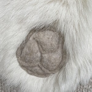 May include: Close-up of a light gray felt applique on a white fur background. The applique is a three-lobed design, possibly a flower or leaf. The fur is long and fluffy, creating a soft texture.