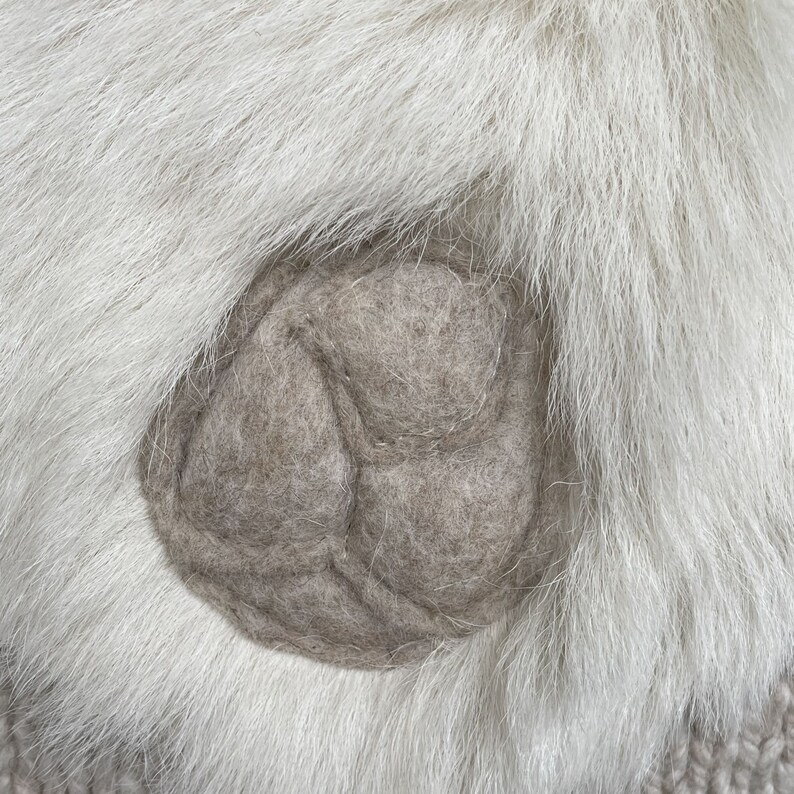 May include: Close-up of a light gray felt applique on a white fur background. The applique is a three-lobed design, possibly a flower or leaf. The fur is long and fluffy, creating a soft texture.
