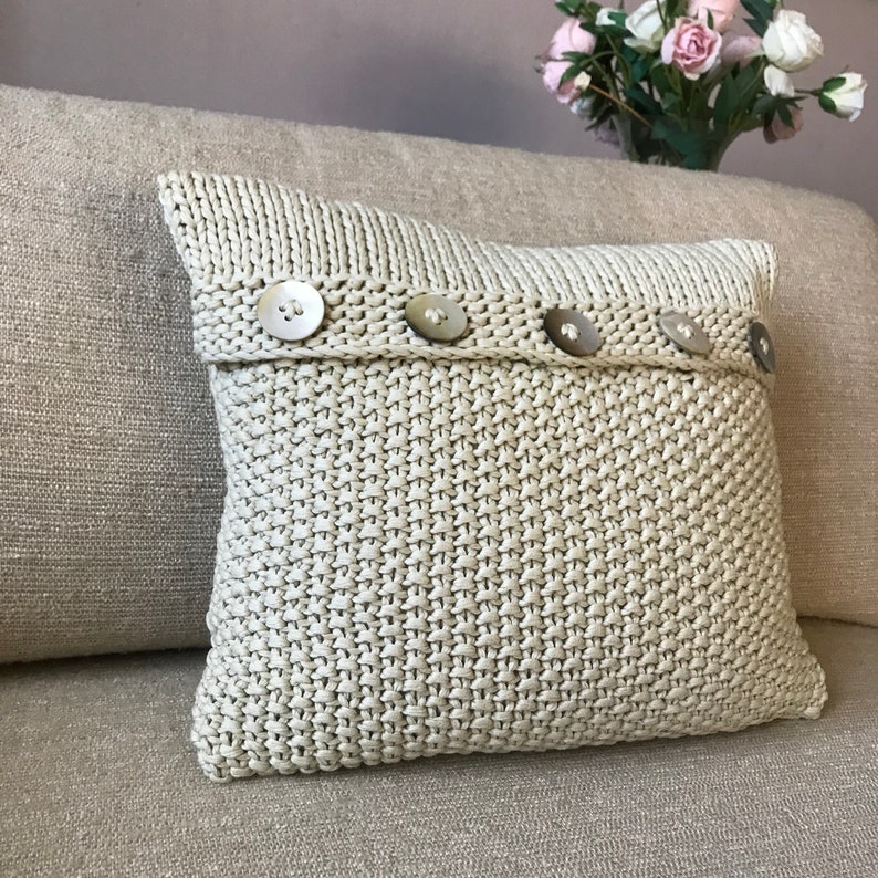 May include: A cream-colored knitted pillow with a row of five decorative buttons.