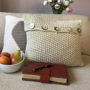 May include: Two knitted throw pillows with button accents on a cream colored couch. A brown leather journal with a pen is on the couch in front of the pillows.