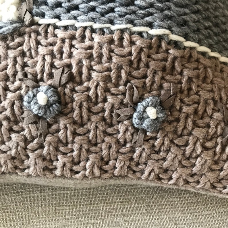 May include: Close-up of a knitted throw pillow featuring a textured weave in shades of taupe and gray.  The pillow is adorned with small, embroidered flower accents in gray and white, adding a touch of rustic charm. The design incorporates a subtle color-blocked pattern, creating visual interest. The pillow's fabric appears soft and cozy, ideal for adding warmth and texture to any living space. The handcrafted details suggest a high-quality, artisan-made product.