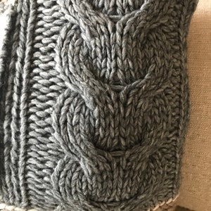 May include: Close-up of a gray cable knit throw pillow. The pillow features a textured, chunky knit with a distinct cable pattern. The gray color is consistent throughout, creating a cozy and warm aesthetic. The knit is dense and tightly woven, suggesting durability and quality. The pillow is ideal for adding texture and warmth to any living space.