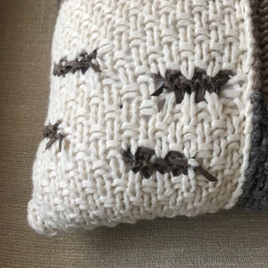 May include: Close-up of a cream-colored knitted throw pillow with brown leather accents. The pillow features a chunky knit texture and decorative brown leather patches stitched onto the cream-colored fabric. The pillow has a casual, cozy aesthetic, suitable for home decor.
