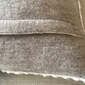 May include: Close-up of a gray wool pillow with a subtle off-white border. The pillow features a textured, nubby surface and a simple, understated design.  The fabric appears soft and thick, suggesting a high-quality, comfortable feel. The neutral color palette makes it a versatile accent piece for various home decor styles. The image highlights the pillow's material and construction details.