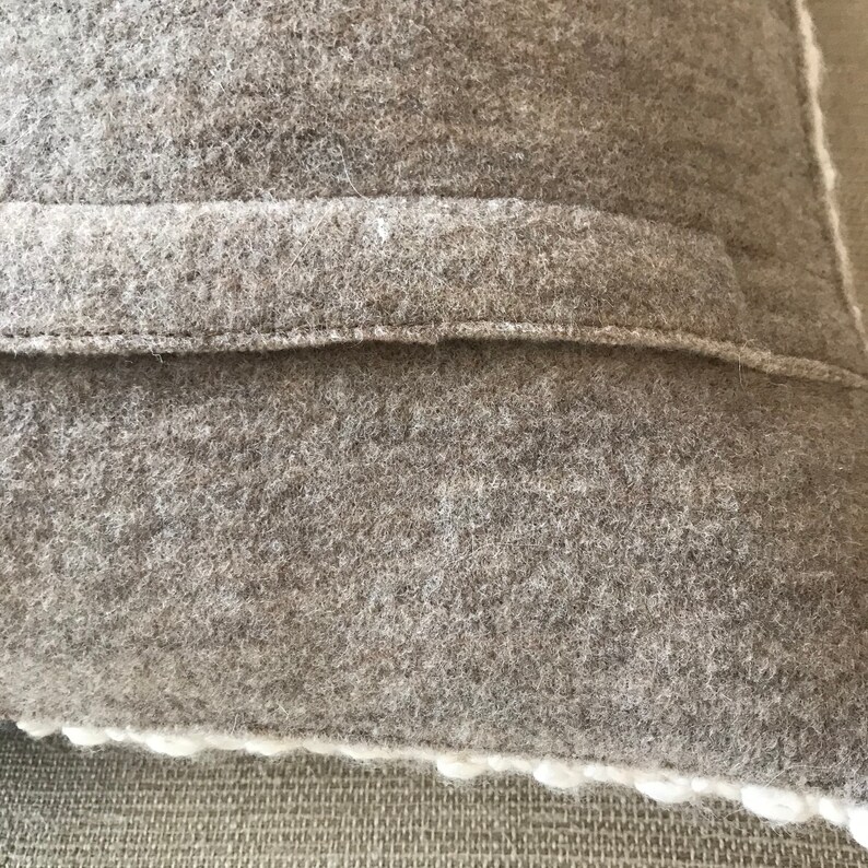 May include: Close-up of a gray wool pillow with a subtle off-white border. The pillow features a textured, nubby surface and a simple, understated design.  The fabric appears soft and thick, suggesting a high-quality, comfortable feel. The neutral color palette makes it a versatile accent piece for various home decor styles. The image highlights the pillow's material and construction details.