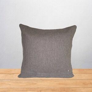 May include: A square, gray decorative pillow with a textured surface. The pillow is made of a soft fabric and has a small, round button on the lower right corner. The pillow is placed on a wooden surface.