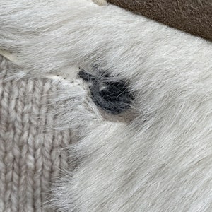 May include: Close-up of a section of a white fur item with a dark gray button. The button is sewn onto the fur, which is adjacent to a light beige knitted fabric. A section of brown leather is visible in the upper right corner.