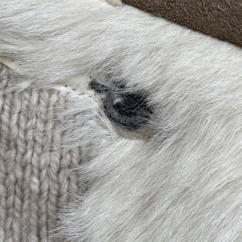 May include: Close-up of a section of a white fur item with a dark gray button. The button is sewn onto the fur, which is adjacent to a light beige knitted fabric. A section of brown leather is visible in the upper right corner.