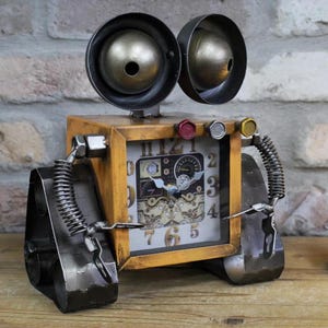 May include: A steampunk robot clock with a yellow metal body. It has large, round, gold-coloured eyes, a clock face, and metal arms and legs. The clock face has a vintage design with Roman numerals and measures approximately 15 cm in height.